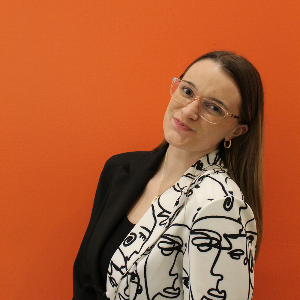 Hailey Danielson wearing a blazer that is half black. The other half is white with abstract faces. She has long brown hair with clear glasses and smiling at the camera. 
