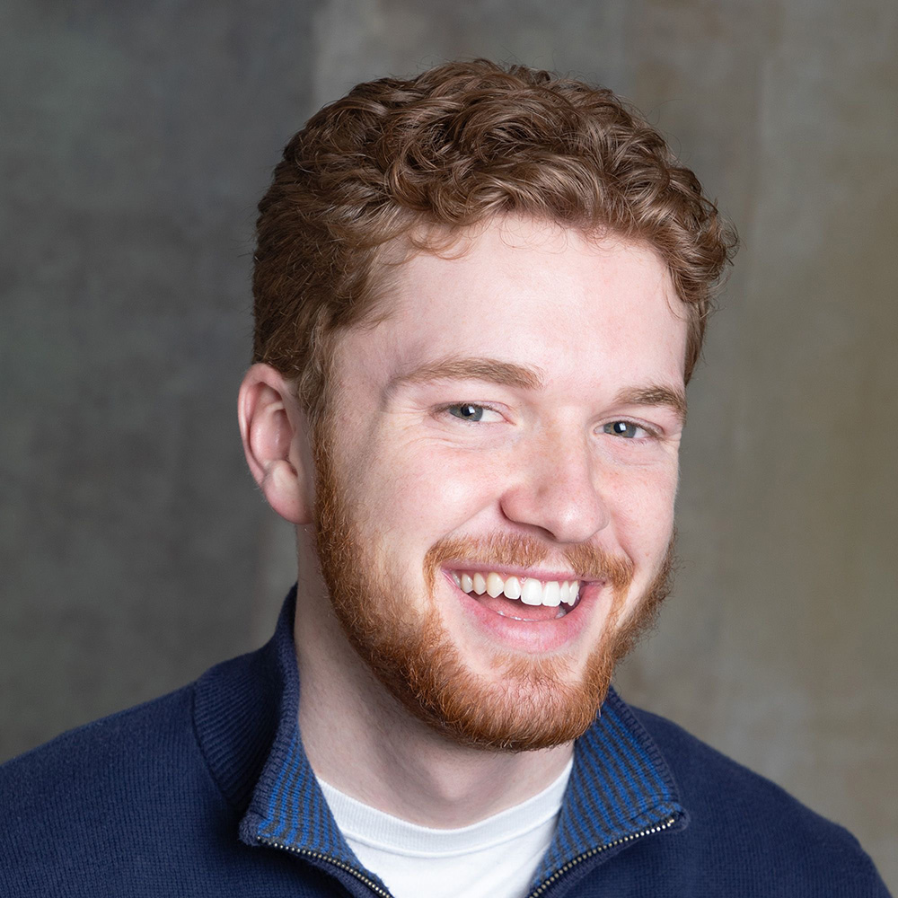 Nicholas Goudsmit wearing a navy blue polo shirt smiling at the camera. He has short reddish-brown hair and a short beard. 